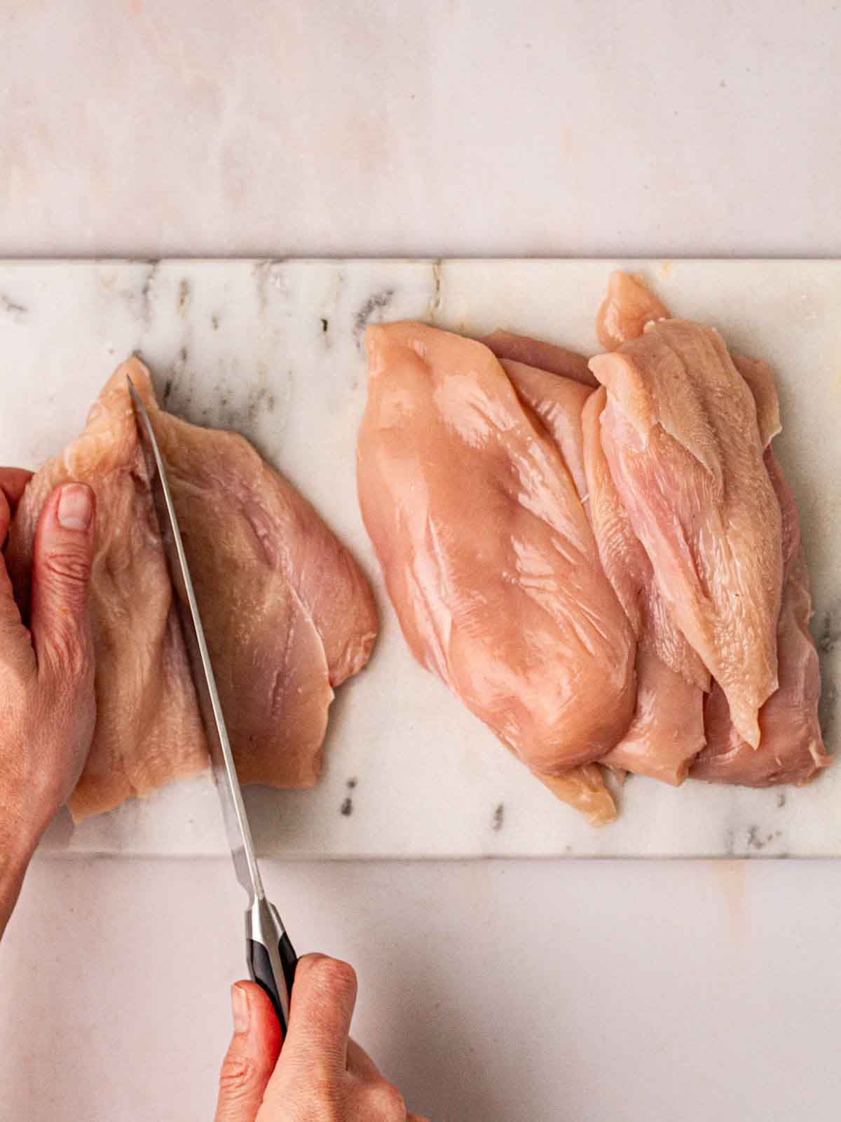 A marble board with two hands slicing chicken breasts in half with a sharp knife.