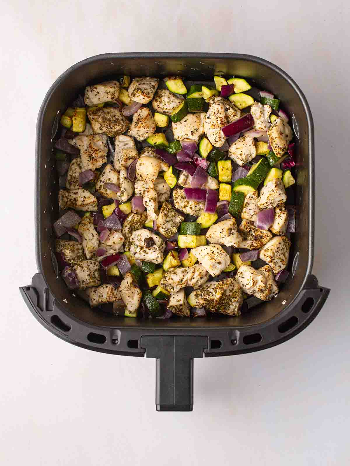 Chunks of chicken breast, red onion, courgette inside an air fryer for Greek Chicken Traybake.