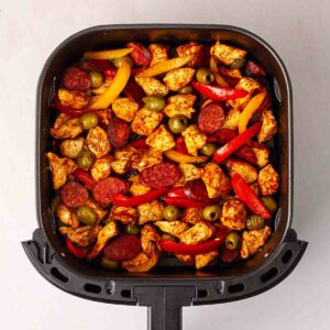 An air fryer with coated chicken, chorizo and veggies in for step 2 in the process for Spanish Chicken.